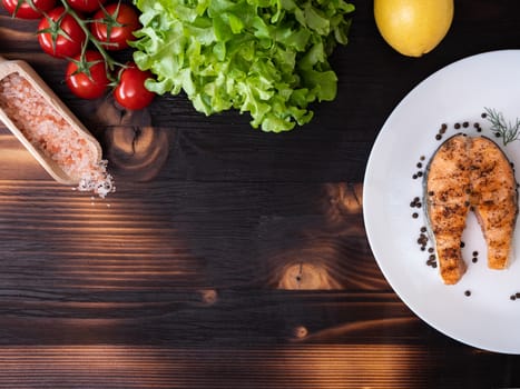 Top view of grilled salmon in a white plate over a wooden background. Salad, cherry tomatoes and lemon next to it