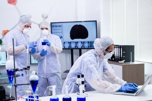 Female scientist working on laptop and colleagues working with reactive blue solution in a modern research laboratory.