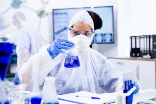 Researcher in protective suit holding a container from which a smoke comes out. Her colleagues are working in the background