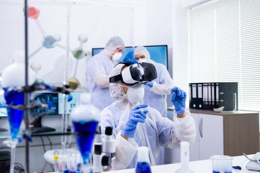 Scientist with virtual reality glasses doing test and her assistants working in the background with reactive blue liquid.