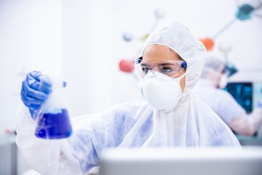 Chemist in goggles and gloves mask holds a container with blue liquid. Modern laboratory