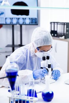 Scientist woman in protection equipment adjusting her microscope. Test tubes with smoking blue solution.