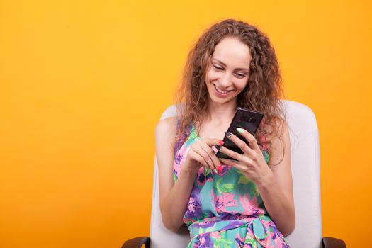 Attractive woman with curly hair smiling using her smartphone in studio over orange background.