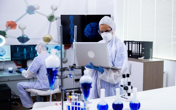 Scientist in modern research laboratory standing and working on laptop wearing protection for smoking blue soltion in test tubes.