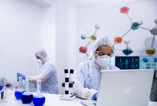 Scientist in protection equipment typing test results on laptop in a modern research laboratory.