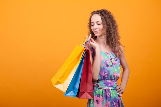Elegance woman in stylish dress looking down on her shopping bags in studio over orange background.