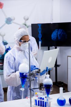 Scientist typing on her computer data from exeperiments with smoking blue solution in test tubes.