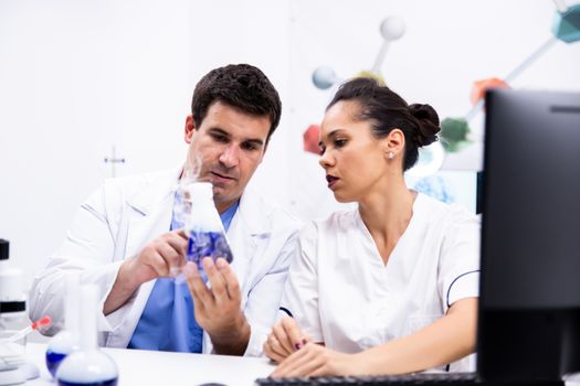 Young scientist in white coat holding a bottle with smoking blue liquid and explaning to his assistant.