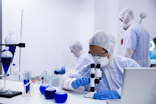 Female scientist looking at a sample of solution through microscope and team of researchers in the background.
