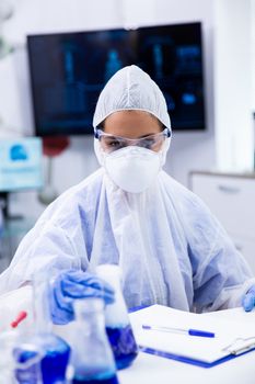 Laboratory dressed in protective suit holds in a container with blue liquid substance. White smoke and goggles