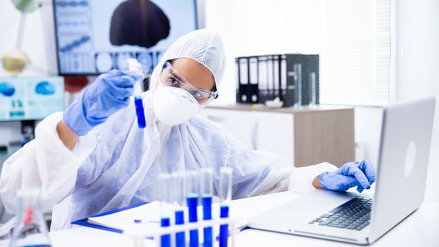 Caucasian scientist analyzes the tube and make some notes on laptop. Blue gloves and goggles