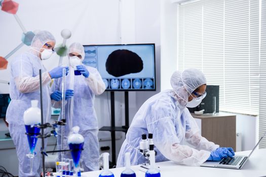 Team of scientists working together with smoking blue solution in modern research laboratory and typing test on laptop.