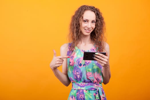 Smiling woman with curly hair pointing with finger at digital device over orange background.