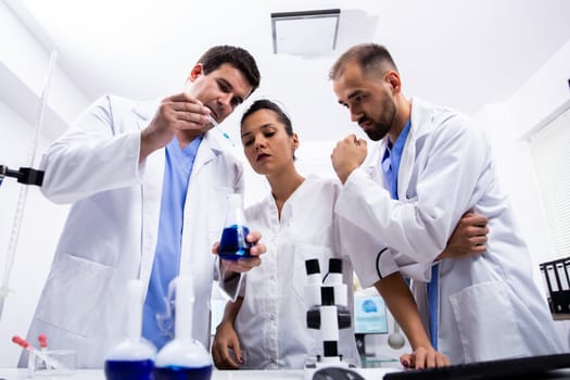 Team of scientist in white coat looking at a sample of smoking blue liquid in research lab.
