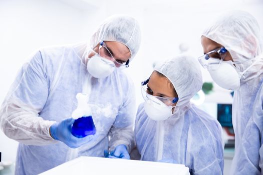 Scientist holds a beaker in his hand and two colleagues follow the reaction. Blue substance and white smoke