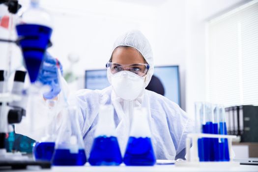 Female scientist in coverall equipment looking at a sample of blue solution in a pipette.