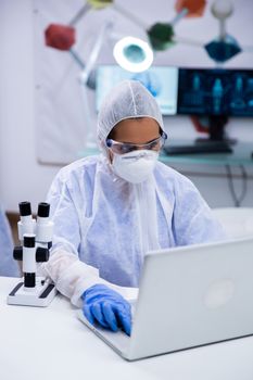 Focused female scientist wearing protection equipment working on her laptop in a modern research laboratory.