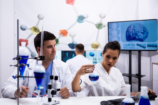 Young scientist wearing a white coat in research laboratory and assistant holding a test tube with blue liquid.