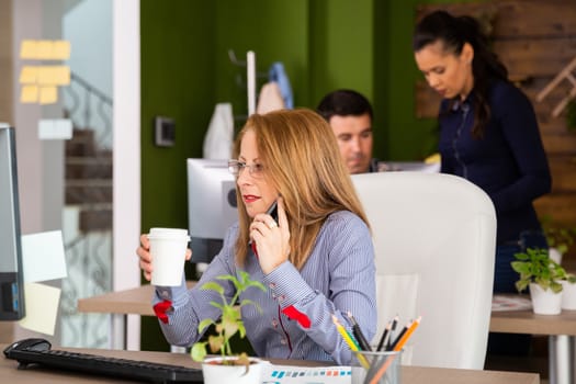 Focused business woman looking into her computer monitor while havine a telephone conversation. Business woman holding cup of coffee.