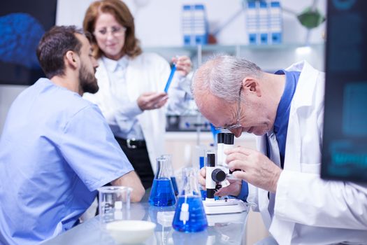Senior scientist doing testing under the microscope in a modern laboratory. Team of scientist working in the background.