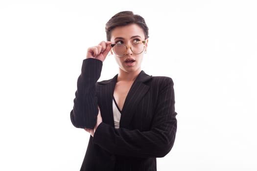 Stylish young fashion model holding her glasses and wearing a black jacket over white background in studio. Cheerful young female.