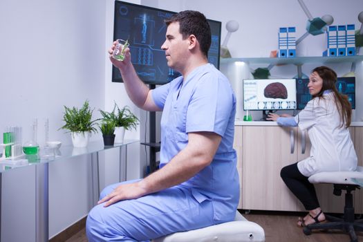 Male scientis holding a sample of green solution in a research lab. Female scientist in the background.