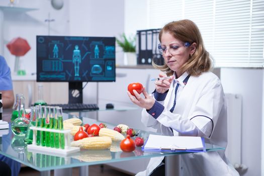 Biologist in white coat in a research lab injecting liquid in a tomato. Test tubes with green liquid.