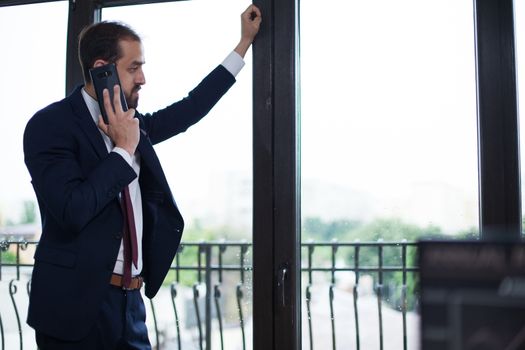 Handsome bearded businessman dressed elegant in his office looking on the window and talking on the phone. Serious entrepreneur.