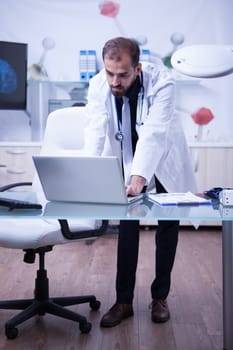 Full lenght portrait of handsome doctor working on his laptop at the hospital cabinet. Doctor in a laboratory.