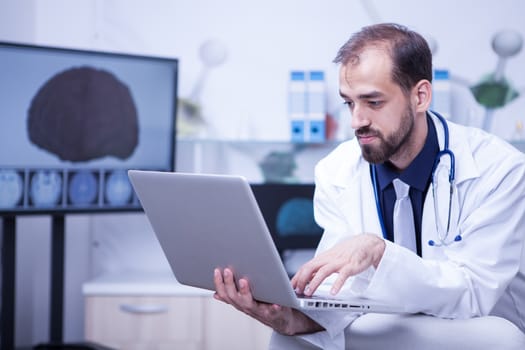 Attractive bearded doctor typing a diagnosticin in the patient file on his laptop. Attractive cardiologist working on laptop.