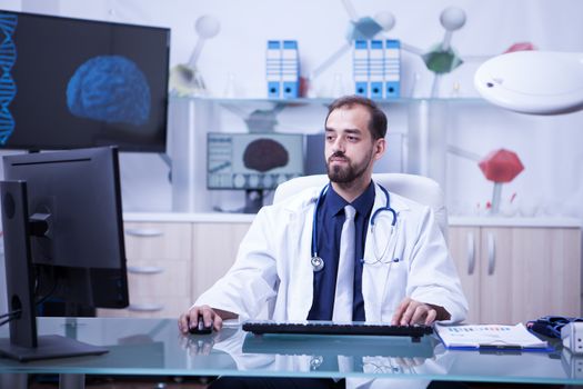 Attractive young doctor doing an online consultation using his computer at the clinic. Young bearded doctor working.