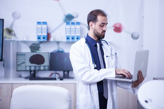Young doctor in his cabinet standing and working on his laptop. Doctor checking the healthcare of patient.