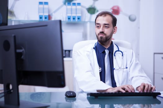 Portrait of doctor working on his computer in cabinet at the hospital. Doctor typing clinic documents.