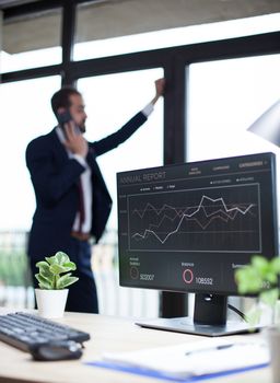 Computer monitor with graphics in young businessman office. Bearded entrepreneur in the background talking on his phone.