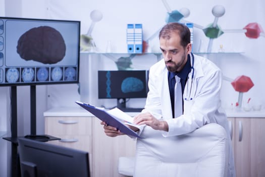 Young and handsome bearded man looking into his monitor and checking his hand notes from clipboard. Doctor in uniform.