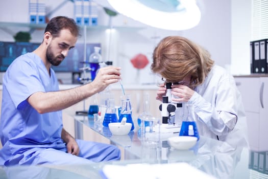 Young caucasian male scientist using a pipette analyzes a liquid to extract molecules in test tube. Female scientist adjusting microscope.