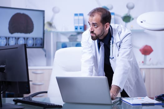 Portrait of doctor working on laptop and looking over his monitor. Doctor on his cabinet. Doctor in a laboratory.