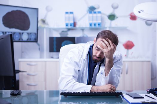 Tired young doctor trying to focus holding his head sitting down in the office. Doctor in laboratory.