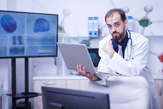 Young bearded doctor looking at the camera and holding a laptop. Doctor using technology.