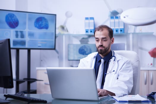 Doctor working on his laptop sitting at his desk in the hospital laboraty. Doctor typing on laptop. Young doctor in white coat.