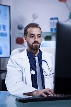 Handsome young bearded doctor typing in his computer in the hospital office. Doctor in white coat with a stethoscope.