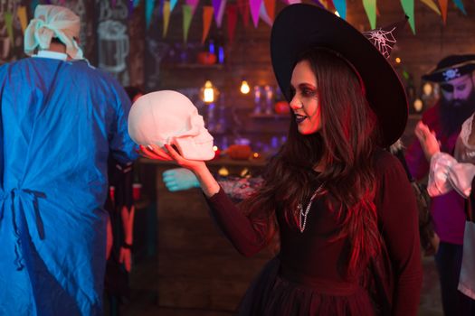 Young attractive witch looking deply into her human skull for halloween party. Celebrating halloween.