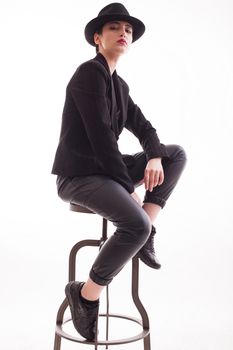 Young fashion model posing with confidence in front of the camera sitting on a chair. Stylish hat.
