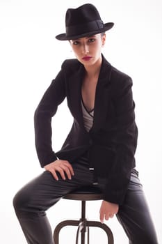 Stylish fashion model posing sitting on a chair wearing a retro hat over a white background in studio. Elegance style.
