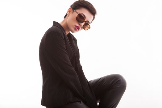 Side view portrait of stylish young fashion model posing in studio sitting on a chair over white background. Stylish sunglasses.