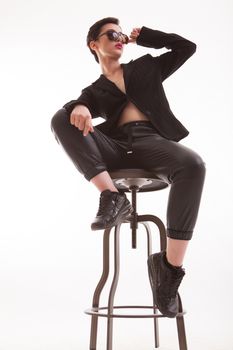 Portrait of attractive young woman sitting on a chair posing in studio over white background. Fashion model holding her sunglasses.