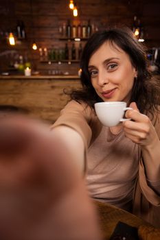 Pretty young woman doing a sef-potrait in a beautiful cafeteria. Beautiful woman smiling at the camera. Beautiful woman holding a cup of mixed coffee.