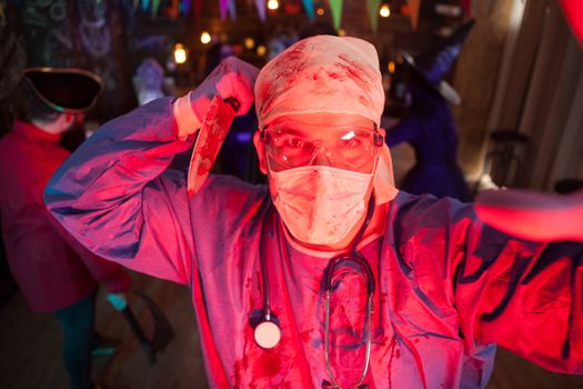 Man holding a knife covered with blood dressed up like a creepy doctor at halloween party. Halloween costume. Friend dressed like monsters in the background.