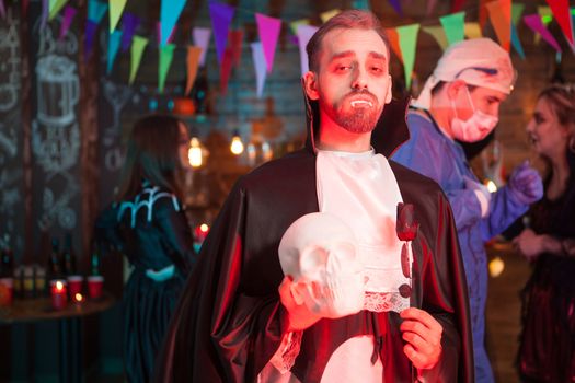 Portrait of Dracula holding a human skull celebrating halloween with his monster friends. Scary doctor covered in blood in the background.