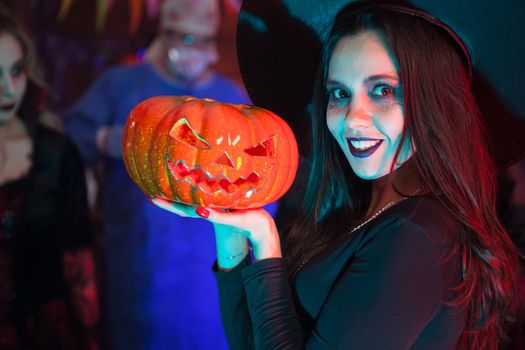 Close up portrait of beautiful woman dressed up like a witch for halloween holding a pumpkin. Halloween event.